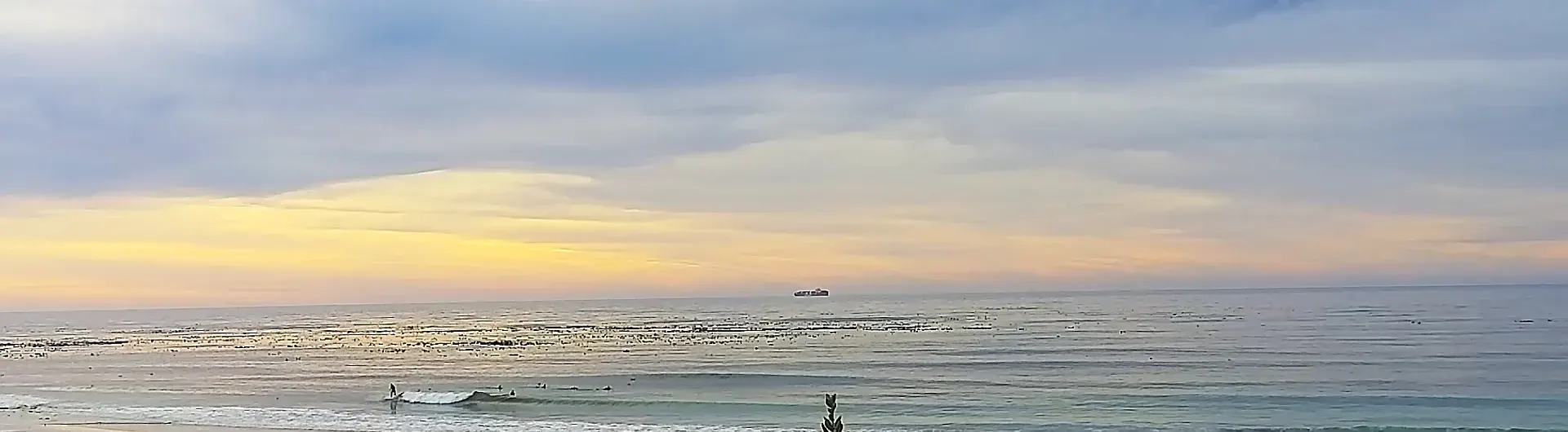 Long Beach from the OceanEye cam at sunset: a surfer on a small wave, a ship on the horizon.